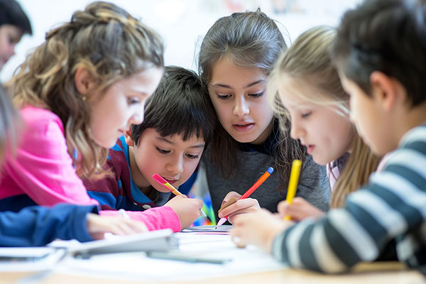 Five kids huddled together at a table with pencils in hand working on a project together