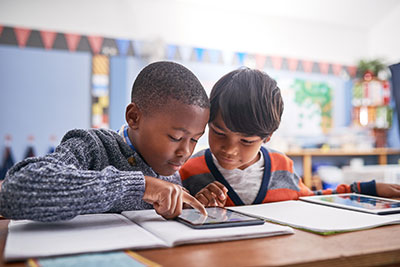 Two children sitting at a long desk staring at content on a tablet